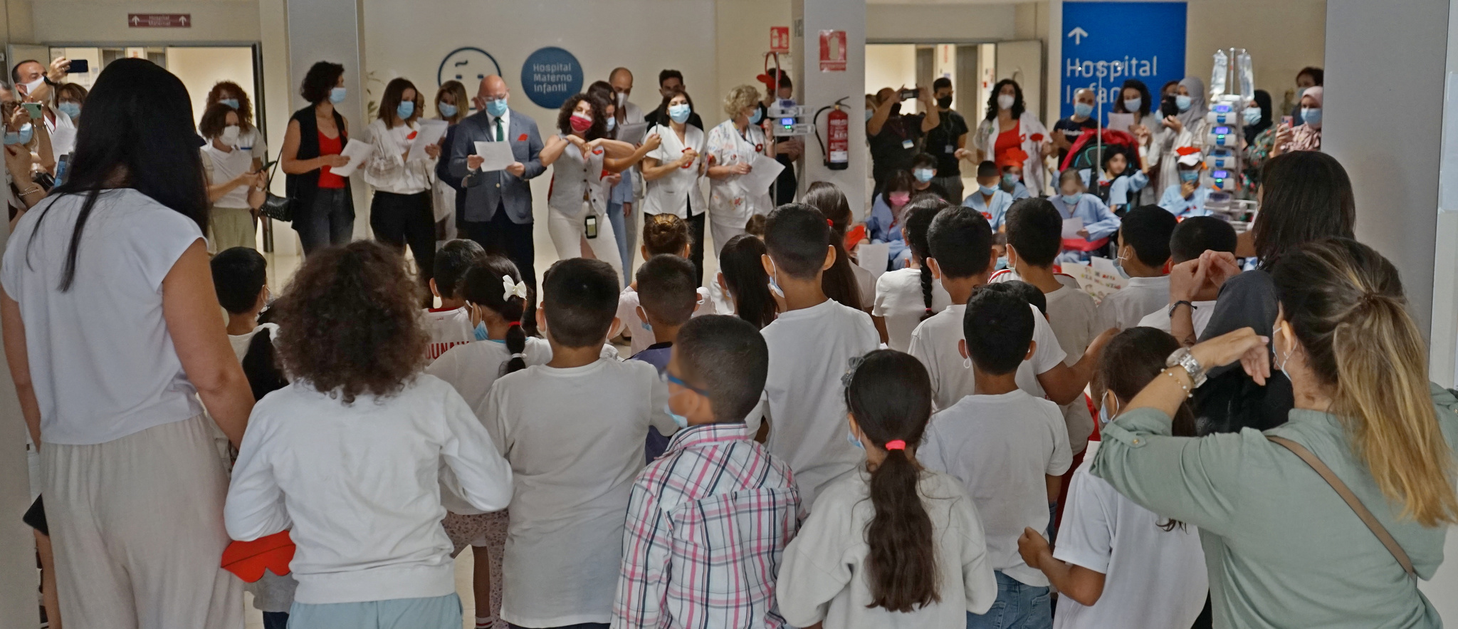 Los niños del CEIP Santa Rosa de Lima (de espaldas) cantan 'Un beso redondo' acompañados de familiares, personal del hospital, voluntarios, el gerente del SMS, Francisco Ponce, y la directora general de Asistencia Sanitaria, Isabel Ayala.